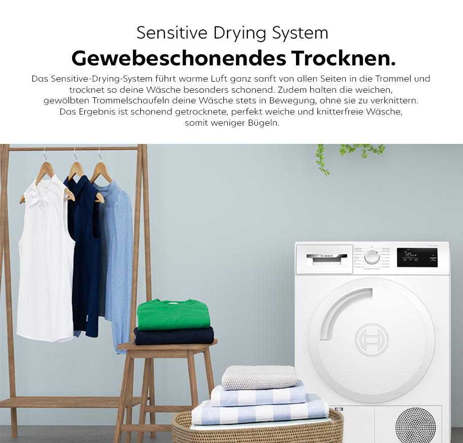 Feature-Banner, Weißer Trockner neben Holzbügelständer mit hängenden weißen, blauen und schwarzen Kleidungsstücken sowie gefalteter Wäsche auf Stuhl und Korb vor grauer Wand.'