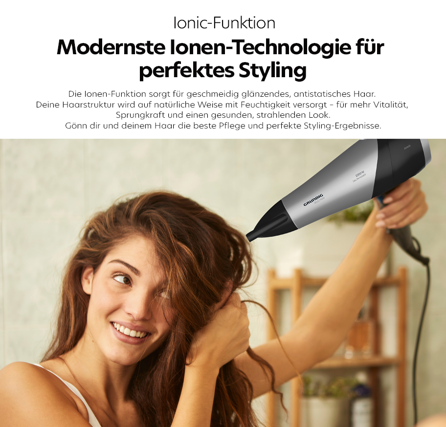 Feature-Banner, Frau mit langen braunen Haaren föhnt ihr Haar mit einem silber-schwarzen Grundig-Haartrockner, im Hintergrund ein Holzregal mit Flaschen und Deko.'