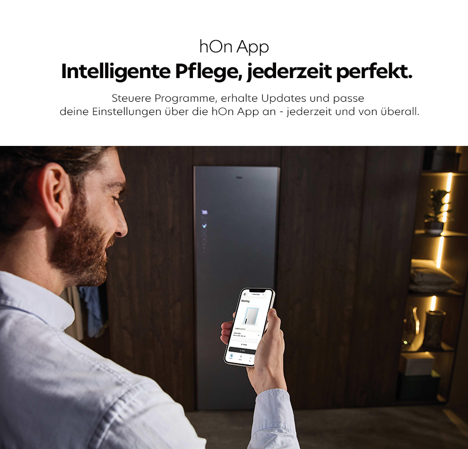 Feature-Banner, Mann hält Smartphone mit geöffneter hOn App vor einen modernen Wäschetrocknerschrank in dunkler Holzumgebung.'