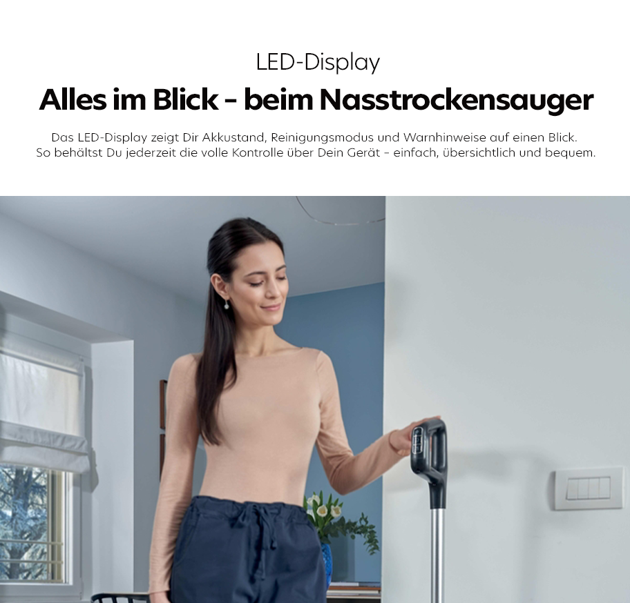 Feature-Banner, Frau mit langem, dunklem Haar trägt beigen Pullover und blaue Hose, hält grauen Nass-Trockensauger in modernem Raum mit Fenster und Lichtschalter.'
