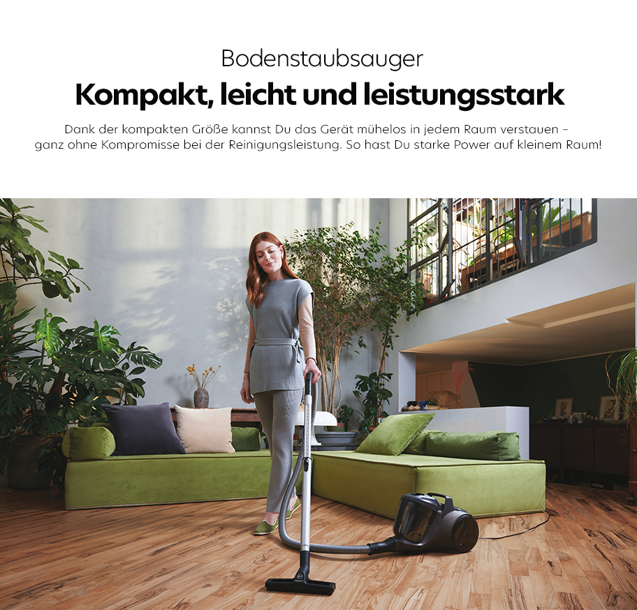 Feature-Banner, Frau in grauer Kleidung steht mit einem kompakten Bodenstaubsauger auf hellem Holzboden vor grünem Ecksofa in modernem Wohnzimmer mit Pflanzen und Treppe.'