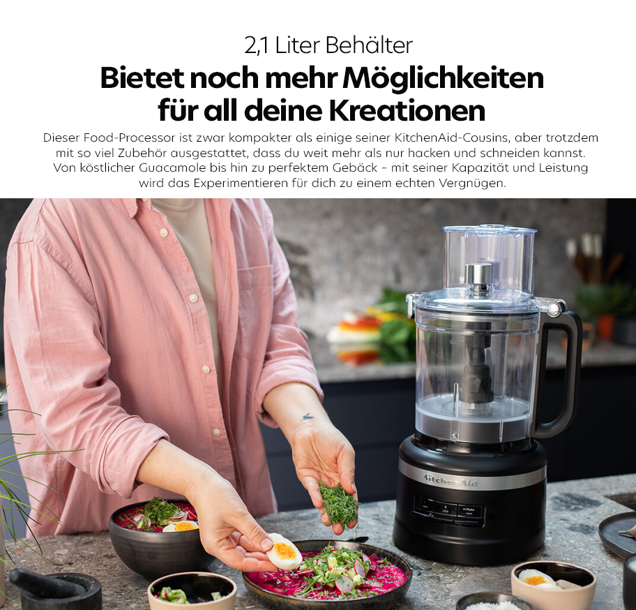 Feature-Banner, Küchenarbeitsplatte mit schwarzem KitchenAid Food-Processor, 2,1-Liter-Behälter, Person in rosa Hemd bereitet Salat mit Kräutern und Ei zu.'