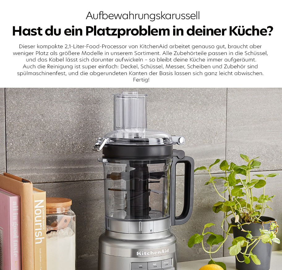Feature-Banner, Kompakter KitchenAid Food Processor aus transparentem Kunststoff mit schwarzem Griff und silberner Basis auf Küchenarbeitsplatte neben Kochbüchern, Glasbehälter und grünem Kräutertopf.'