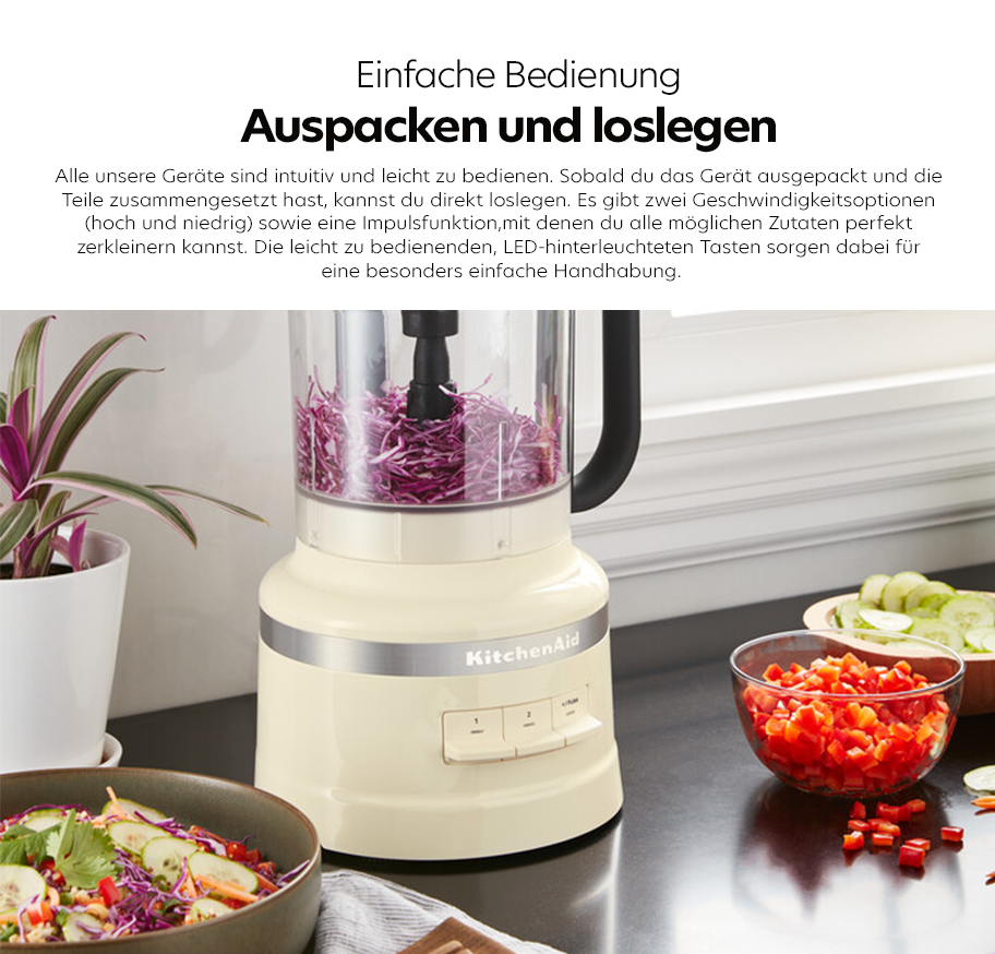 Feature-Banner, Küchenmaschine von KitchenAid mit durchsichtiger Rührschüssel, gefüllt mit geschnittenem Rotkohl, auf schwarzer Arbeitsplatte neben Schalen mit geschnittenem Gemüse und Salat.'