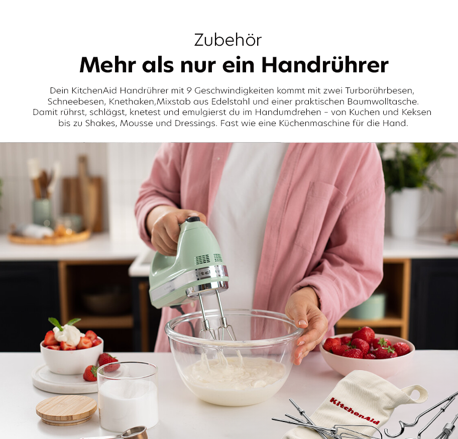 Feature-Banner, Person in rosa Jacke hält grünen KitchenAid Handrührer über Glasschüssel mit weißer Masse, daneben Schüssel mit Erdbeeren, Glas mit Milch und KüchenAid Baumwolltasche mit Zubehör auf weißer Arbeitsfläche.'
