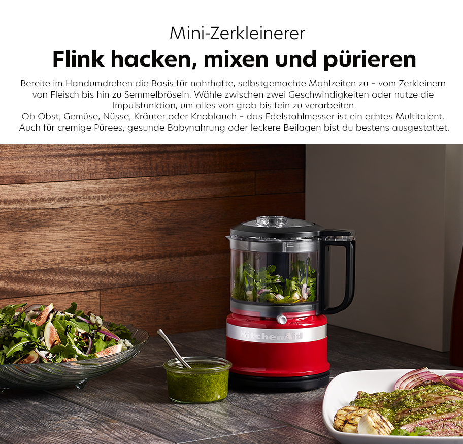 Feature-Banner, Roter Mini-Zerkleinerer von KitchenAid mit Edelstahlmesser, daneben Schüssel mit Salat, Glas mit grüner Sauce und Teller mit gewürztem Fleisch.'